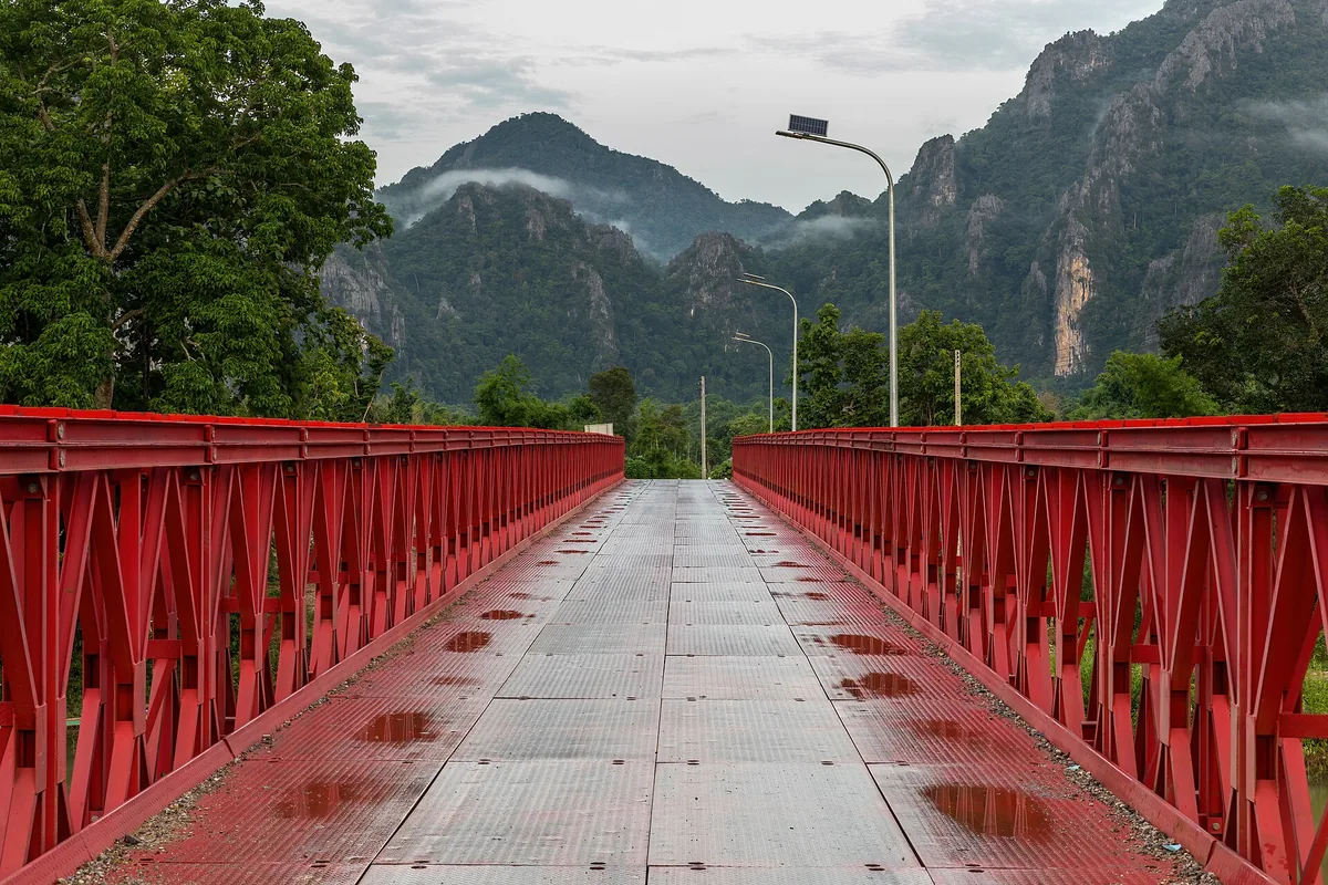 A red steel bridge crossing the Nam Song river at Vang Vieng, limestone peaks in cloud behind it.