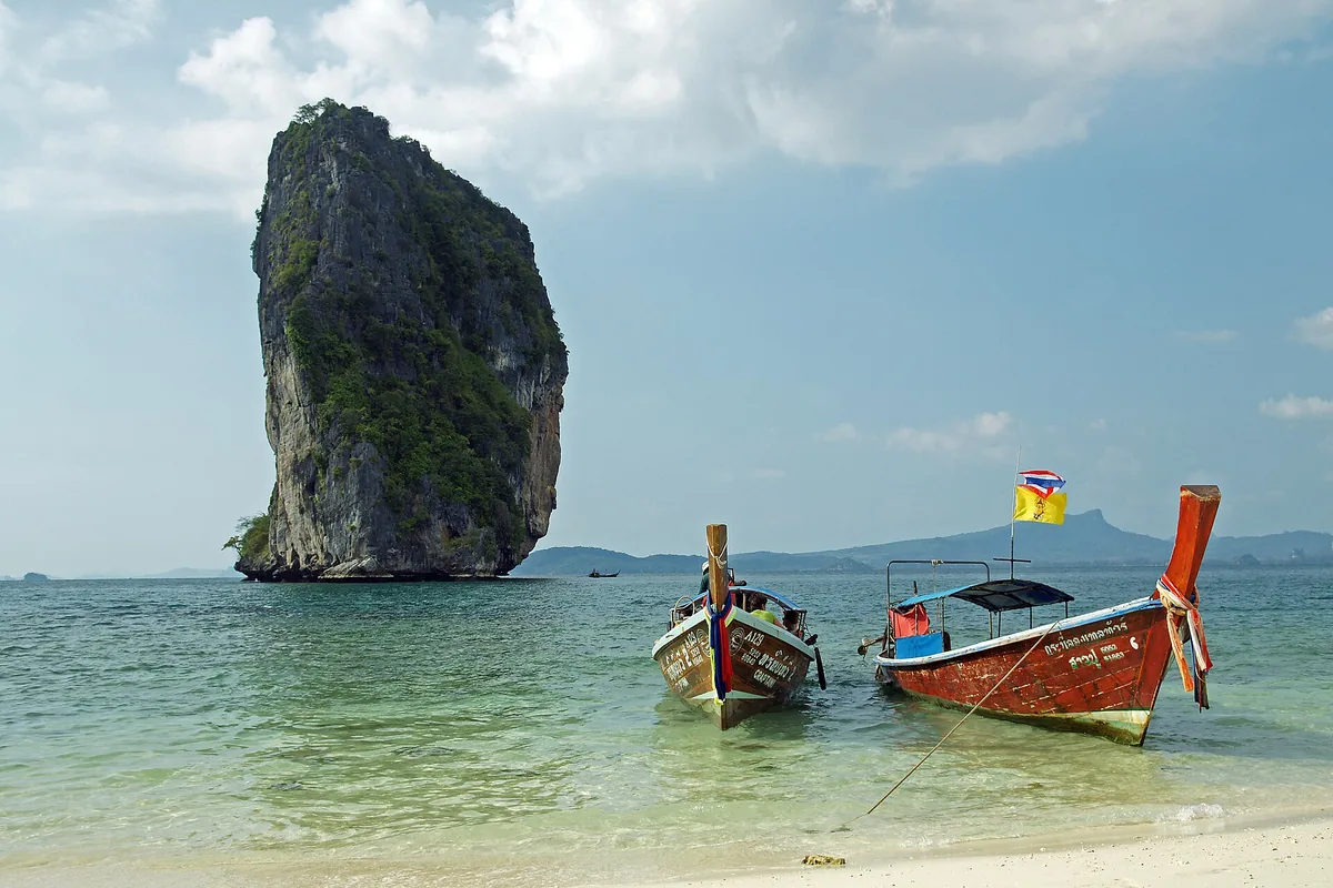 A single limestone stack offshore at Koh Poda, longtail boats moored in clear turquoise shallows.