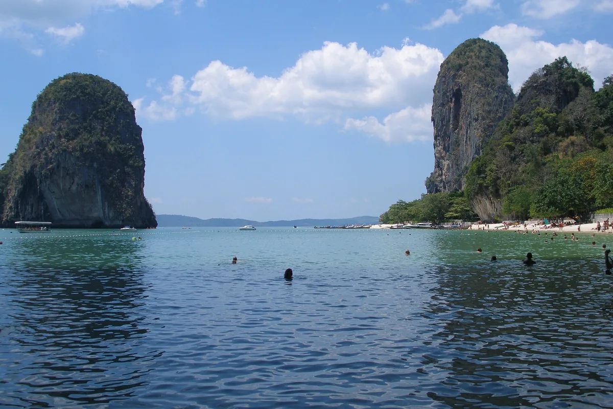 Two limestone towers standing straight out of shallow green water at Railay, with swimmers in the foreground.