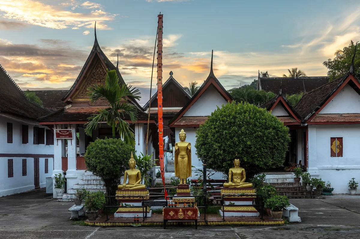 Three gilded Buddha images in the courtyard of Wat Mai, Luang Prabang, under a sunset sky.